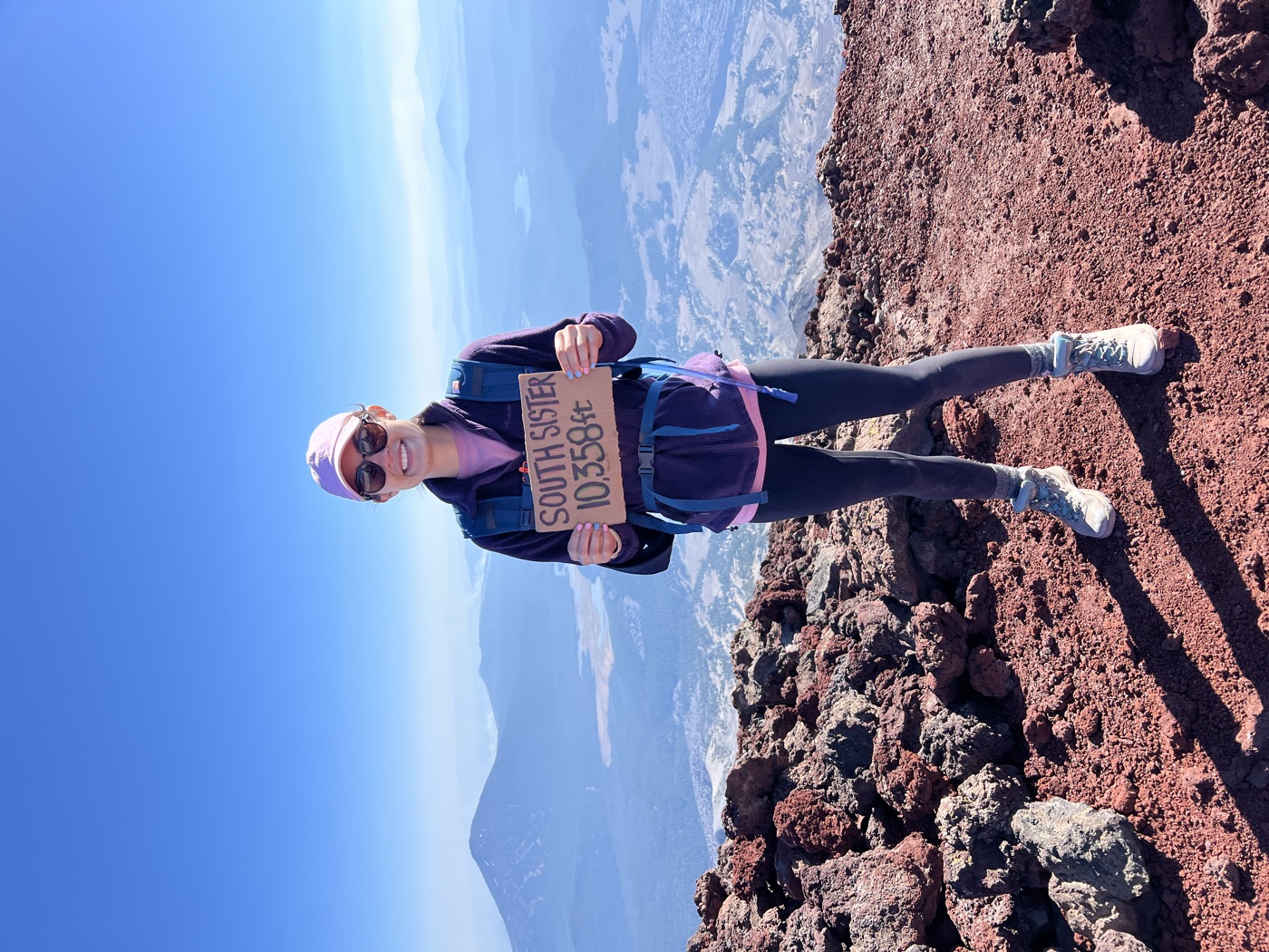 Lilly on the red volcanic rock at the summit of South Sister, holding a cardboard sign reading South Sister 10,358 ft, with Oregon's Cascades hazy blue behind.